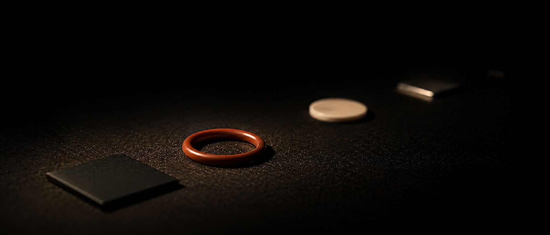Four vacuum samples on a dark bench, a black coupon, an orange elastomer O-ring, a pale ceramic disc and a steel plate, each one further from the light than the last until the darkness takes them.